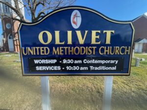 Blue sign with white text for olivet united methodist church listing worship service times, with a tree and church building in the background.
