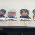 Hockey sticks mounted on a wall, each featuring a photo of a different child's face with helmets, labeled with encouragement signs like 